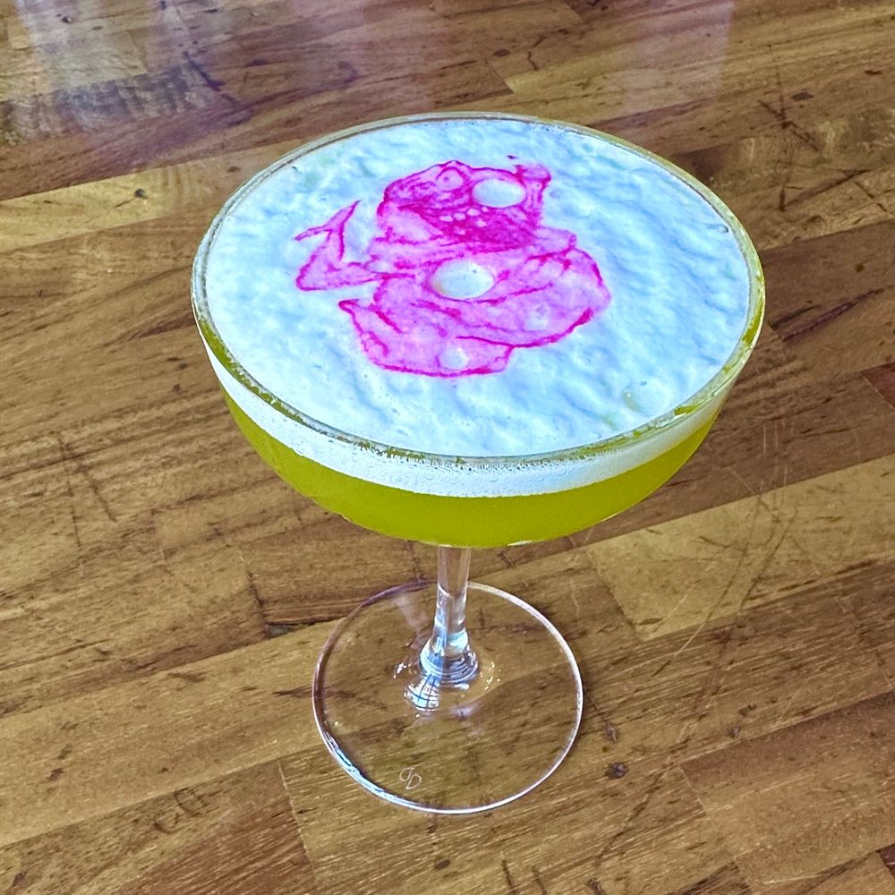 A bright green cocktail in a tall coupe glass with a pink imprint of a Ghostbuster character in the foam on the top of the liquid.