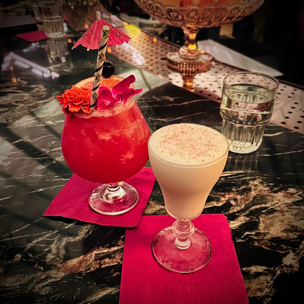 Two cocktails on a marble counter. On the left a tropical red cocktail with a paper umbrella. On the right a creamy yellow cocktail in a tall fluted glass. The glasses sit on red paper napkins.