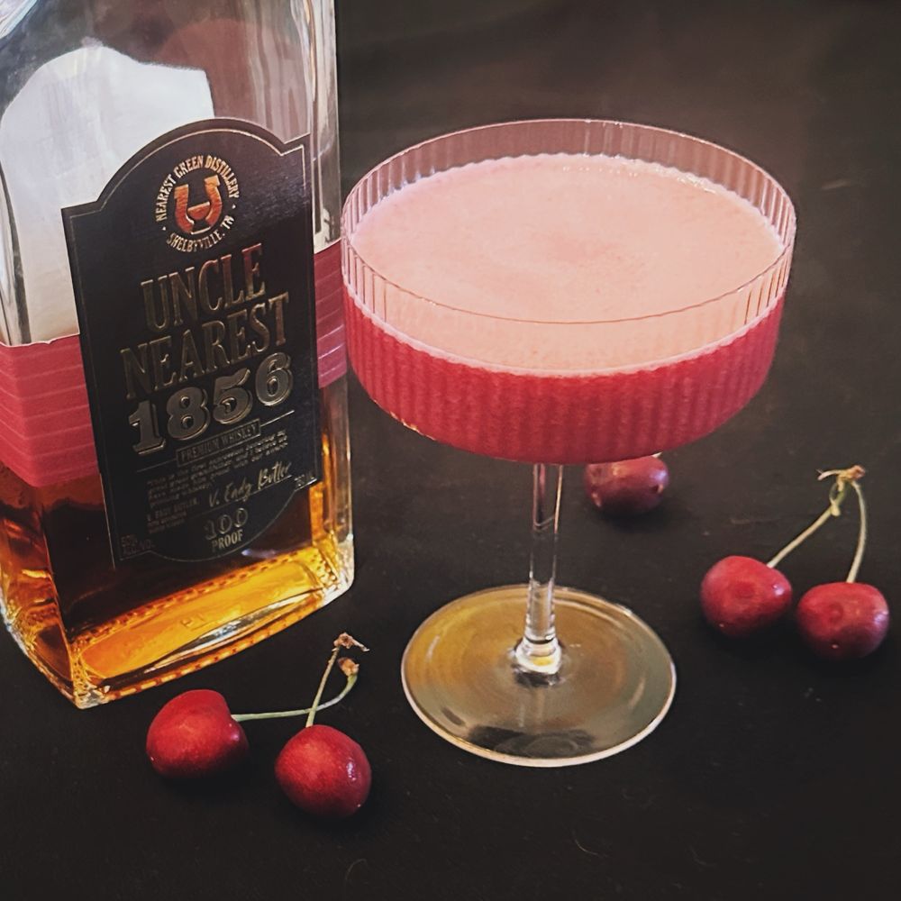 A red cocktail in a coupe glass surrounded by cherries and a bottle of whiskey
