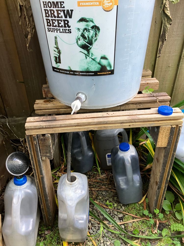 A 20+ litre plastic home brew barrel sits atop a wooden frame. A tap on the front of the barrel has a plastic tube going down into an empty and dark stained 3litre milk bottle. Other milk bottles stand around it, containing some dark foul liquid. Definitely not milk. This is a bottling operation