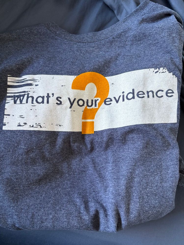 Grey t shirt with a white text box saying “what’s your evidence” with a large orange question mark. 