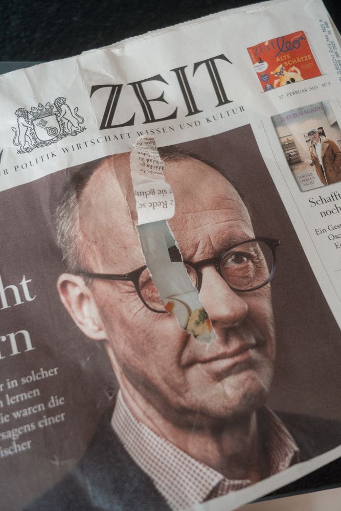 Eine zerrissene Zeitung mit einem Nahaufnahmeporträt von Friedrich Merz. Die Zeitung trägt den Titel "ZEIT" und enthält deutschen Text. Das Bild ist teilweise durch einen Riss im Papier verdeckt, der einige bunte Reste darunter enthüllt.