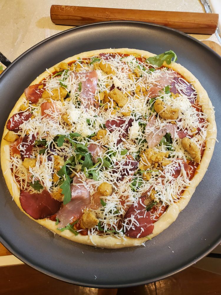 A picture of a homemade pizza on a pizza pan before it goes in the oven. The toppings are cheese, arugula, Prosciutto, and chicken.