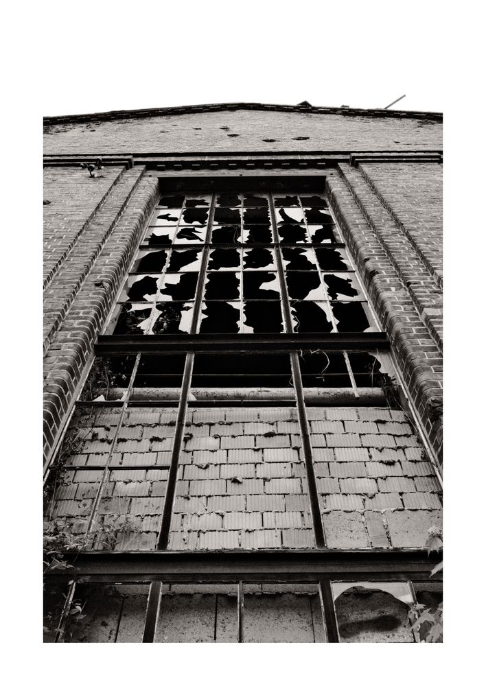 Schwarzweißfoto einer hohen, zerbrochenen Fensterfront eines alten Backsteingebäudes, aufgenommen aus der Froschperspektive. Die Glasscheiben sind größtenteils zerstört, und die Fassade zeigt deutliche Spuren von Verfall.