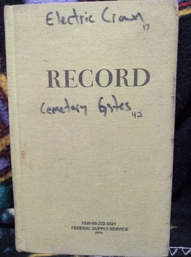 Hardcover green government-issue notebook with handwritten story titles "ELECTRIC CROWN" and "CEMETERY GATES"