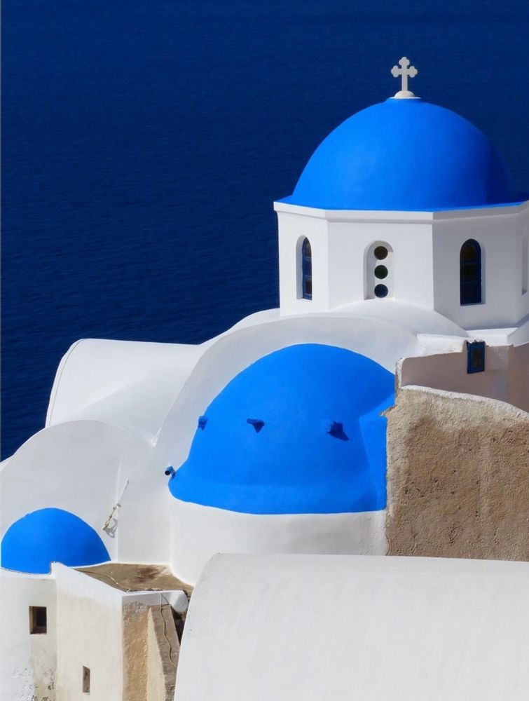 This is a picture of three domes topping whitewashed buildings in Oia, Santorini. The buildings are on a steep cliff, dropping off into a sea that looks navy blue. The domes are a very deep matte turquoise paint, making a great contrast with the white structures. The largest domed roof has a white crucifix on top. 
