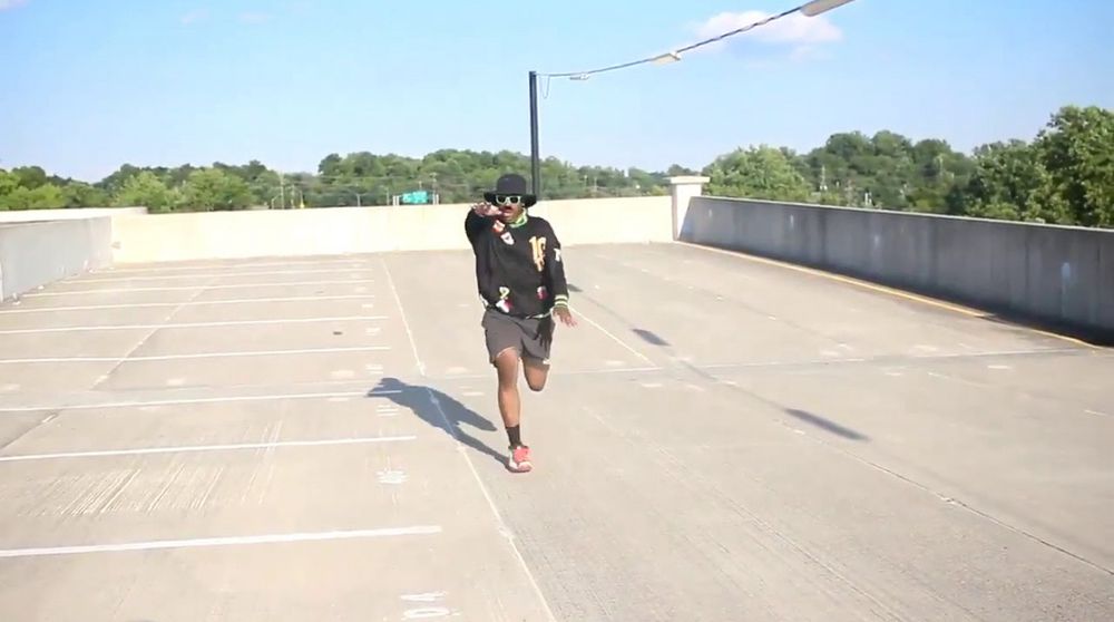 Man in a hat running towards the camera with his hand extended pointing at the person behind the camera 