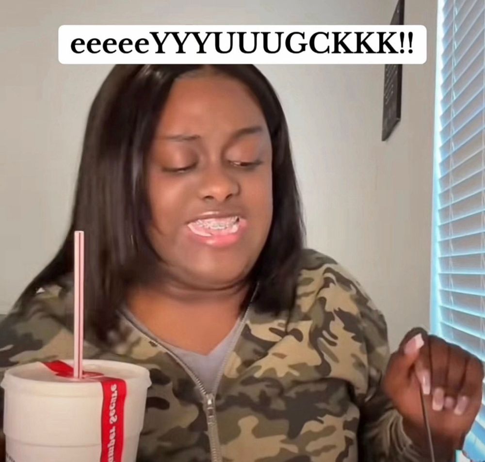 A Black lady in a camo hoodie sitting at a table saying EEEEYUCK about the mac and cheese she just sampled