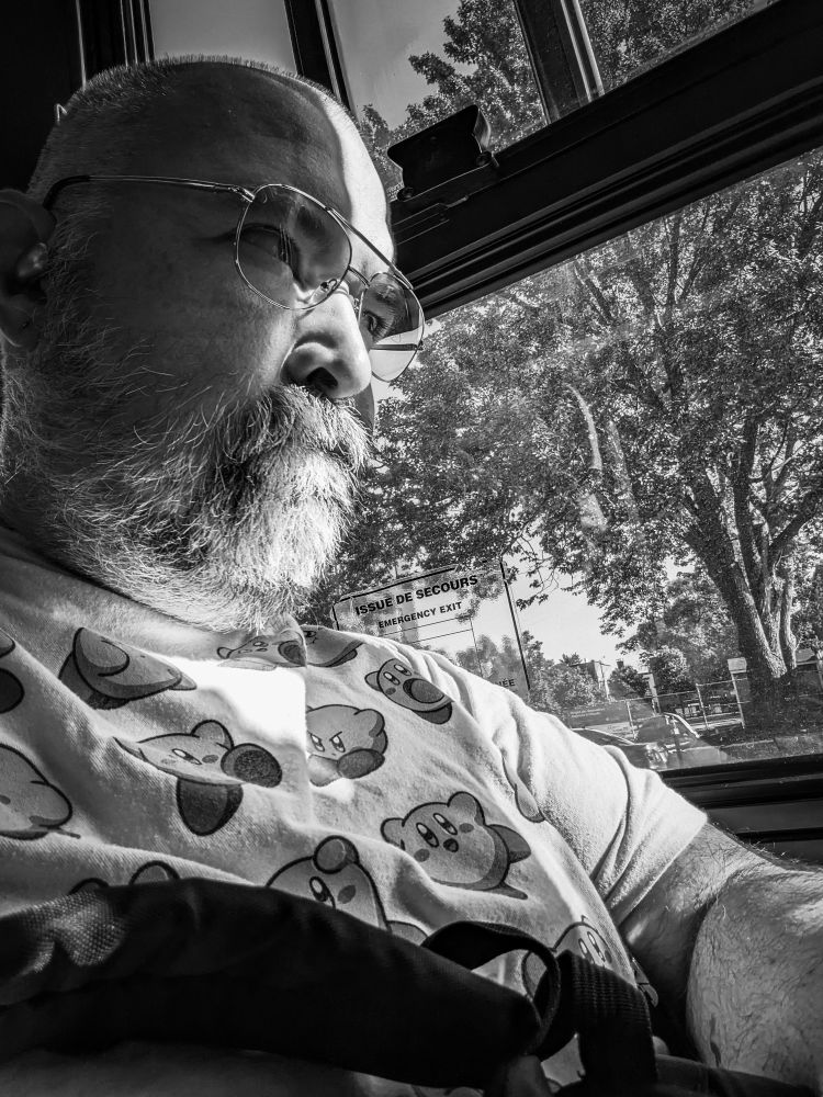 A black and white photo of a fat man glowering out of a bus window with a bright step of sunlight on his face. His expression is countered by an adorable Kirby t shirt. 