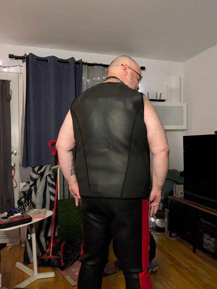 A fat man wearing a neoprene black and blue vest, black harness with pride colored studs, a black neoprene tank top  and red and black neoprene pants from the back