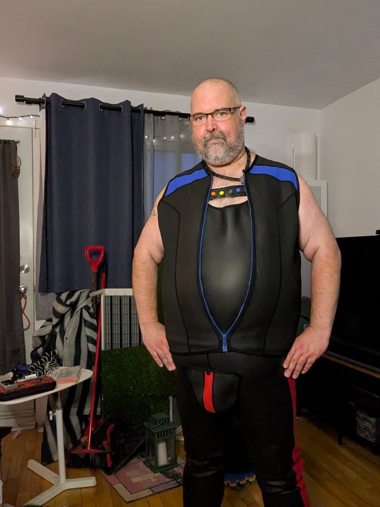 A fat man wearing a neoprene black and blue vest, black harness with pride colored studs, a black neoprene tank top  and red and black neoprene pants
