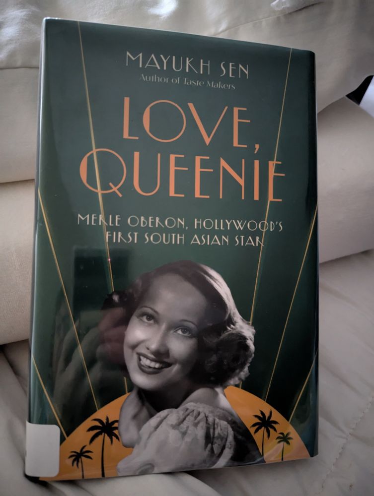 Book cover with the main title "Love, Queenie" in large orange letters on a dark green background. The bottom half of the cover is a photo of actress Merle Oberon, from shoulders up, smiling.