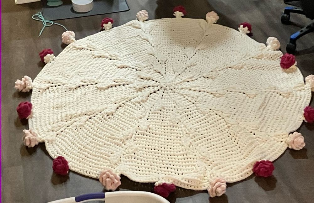 Large circular white crochet blanket on the floor, with pink roses attached to its edges. 