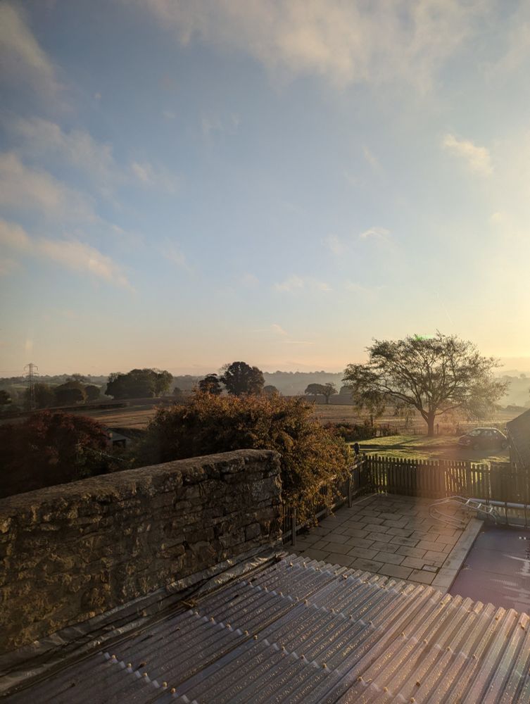 View out of the bedroom window looking over the misty fields. The sky is blue with a few white clouds. The sun is streaming into the side of the view.