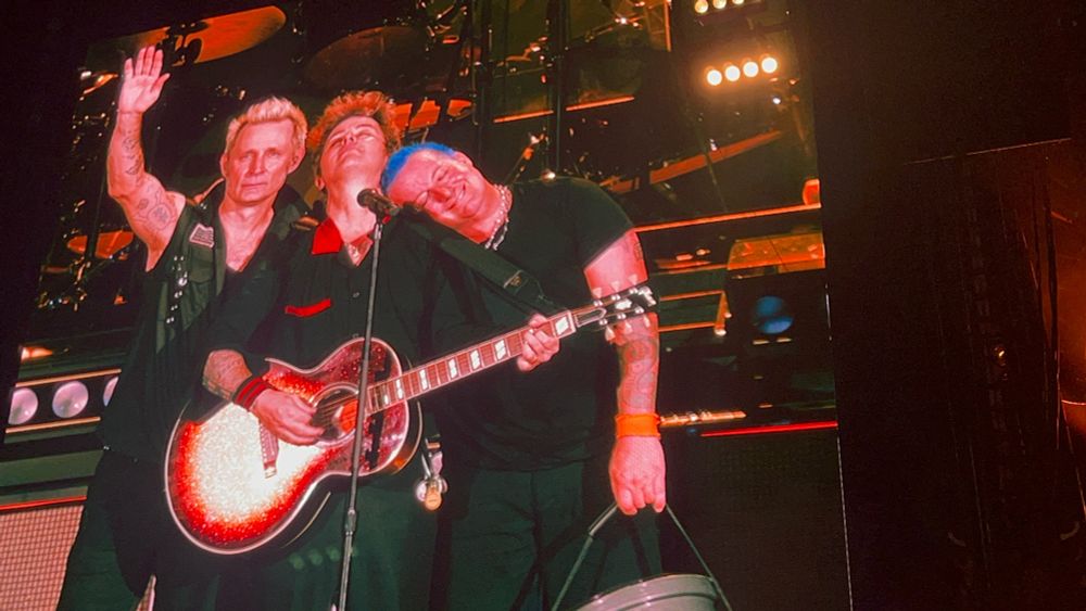 The members of Green Day stand should to shoulder behind the microphone. 