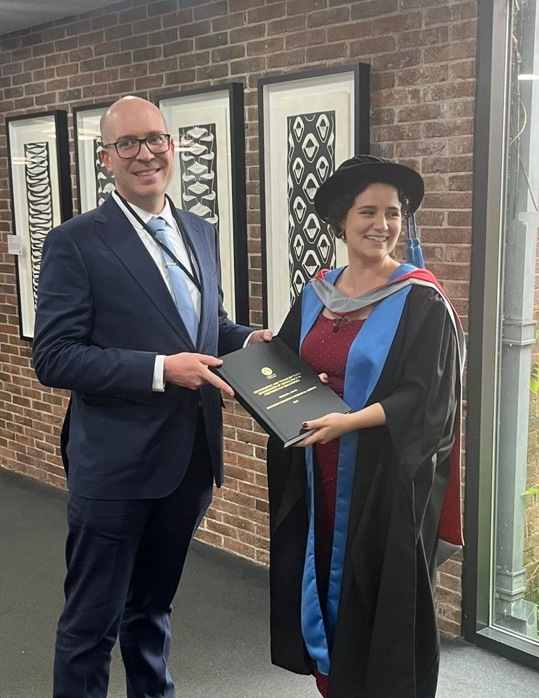 Rebekah and supervisor holding PhD thesis. Rebekah is in graduation robes and smiling at someone off camera.