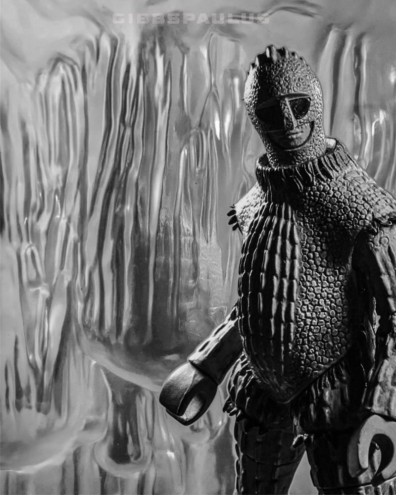 A giant reptilian humanoid stands inside a cave of ice, its comrade frozen inside the wall.