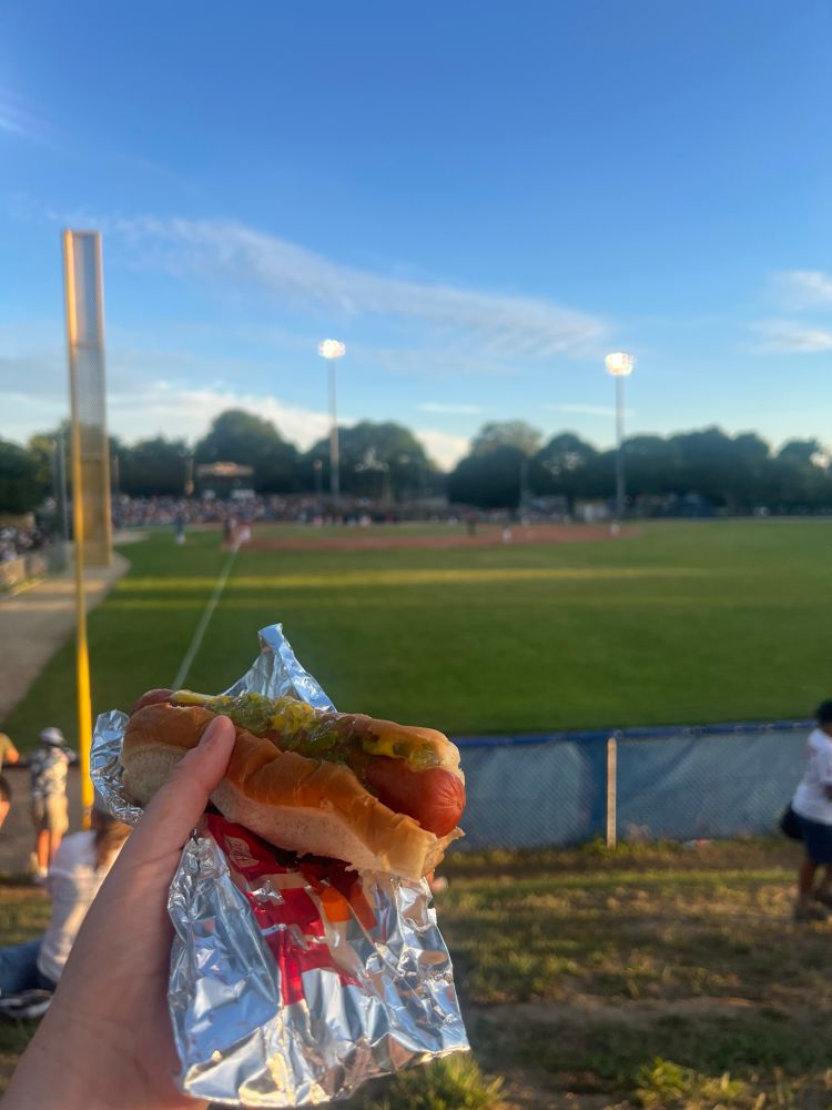 A hotdog with yellow mustard and relish is held in the foreground along with the foil wrapper in which it was originally wrapped. A baseball diamond is visible but blurry in the background. It is a beautiful golden hour with blue sky and bright cloud.