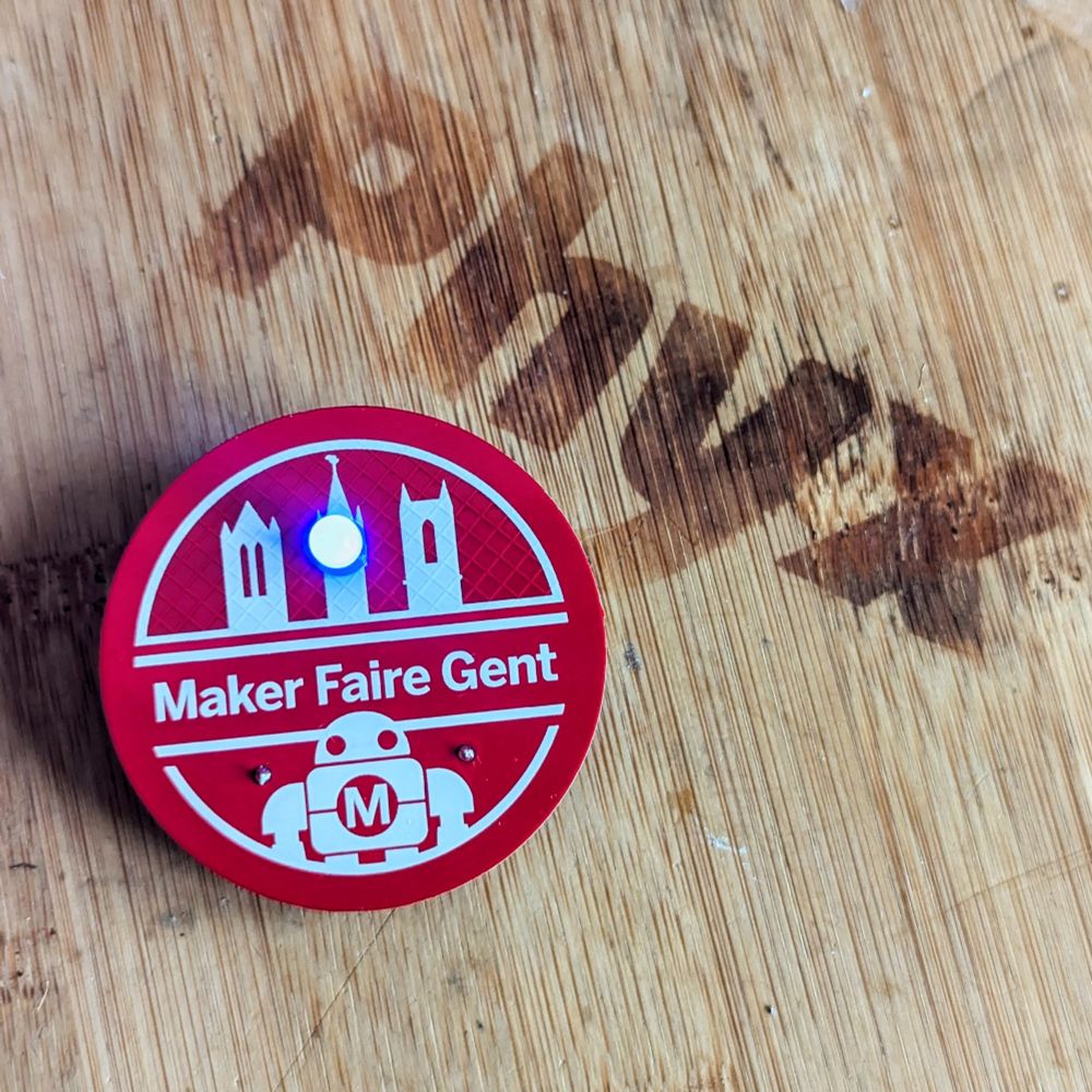 A 7cm round red PCB with white solder mask in the likeness of the Maker Faire Gent logo with a single lit blue LED. 
