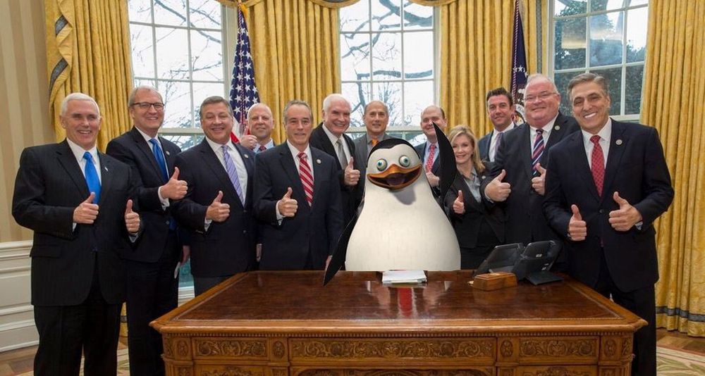 Trump as a giant penguin signing an executive order while being given a thumbs up by his team.