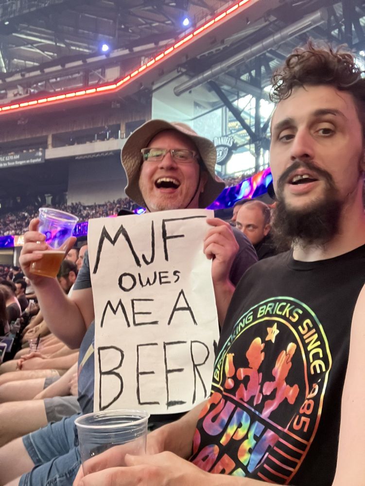Myself and a friend in our seats at AEW All In Texas. I am holding a sign that says "MJF owes me a beer!"