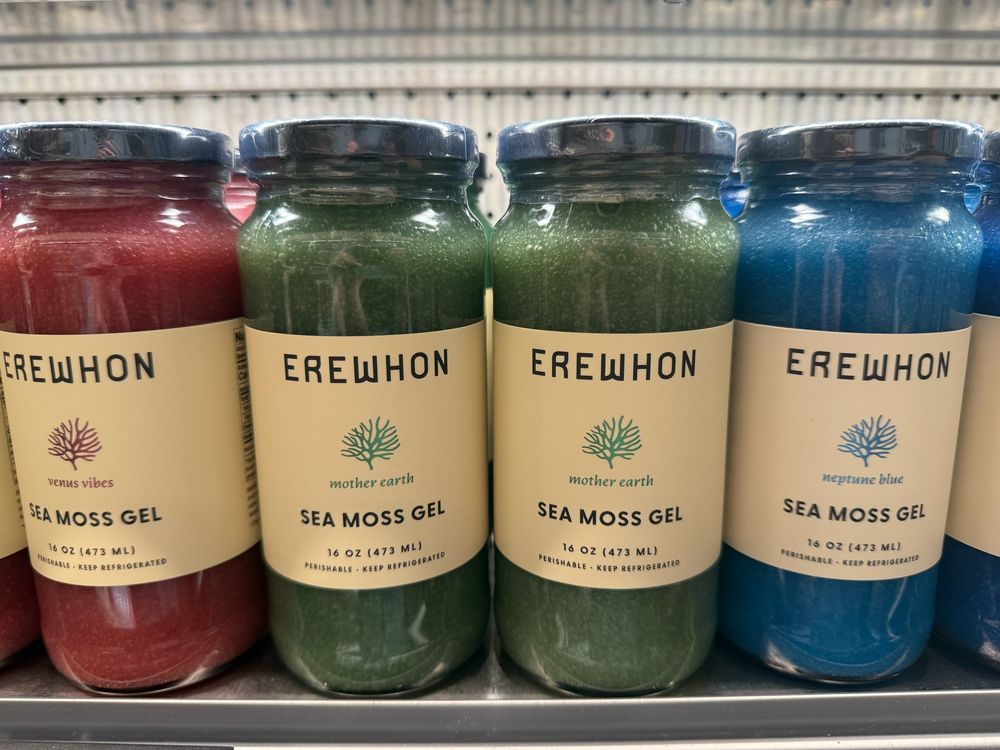 A row of see-through glass jars labeled Sea Moss Gel in a rainbow of colors (red, green, blue) on the shelf at Erewhon in LA.