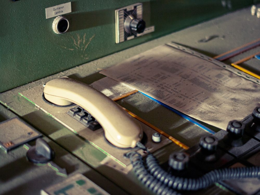 The photo shows a close-up view of an old control panel covered in dust. In the foreground, there is a beige telephone handset resting on its cradle, attached by a coiled black cord. Several push buttons are visible beneath the handset. To the right, a sheet of paper with printed text and numbers lies on the surface, slightly yellowed with age. The panel itself has a dull green finish and is fitted with knobs, switches, and labels. One label, above a circular button, reads “Quittieren Lambda.” The equipment appears worn and unused, suggesting it belongs to an abandoned or decommissioned facility.