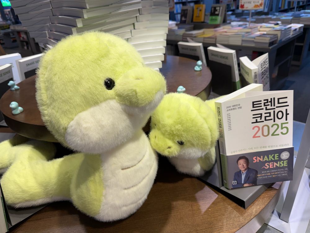 서점 테이블에 [트렌드 코리아 2025]라는 제목의 책이 큰, 연초록 뱀 봉제인형 두 마리와 전시되어 있고, 그 뒤로 보이는 책탑 주면으로 작은 하늘색 뱀 피겨들이 원형으로 놓여있다.