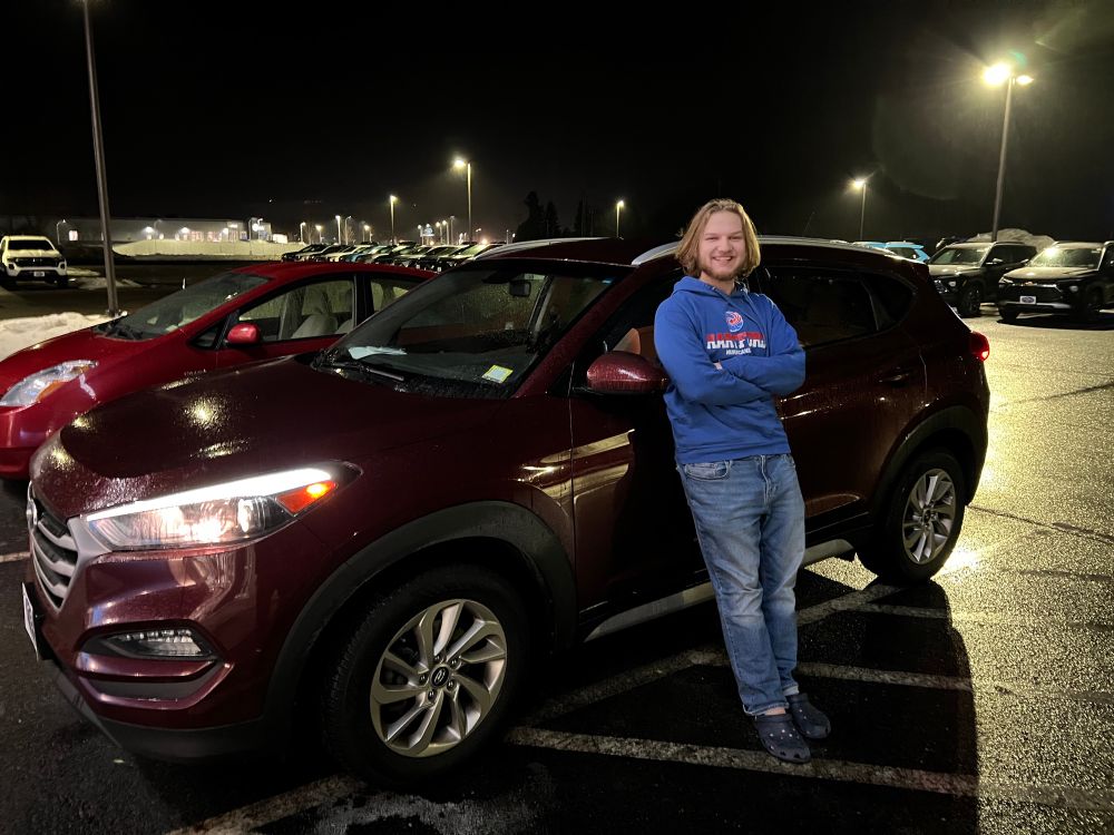 Smiling teen in sweatshirt and jeans leaning against a red used car