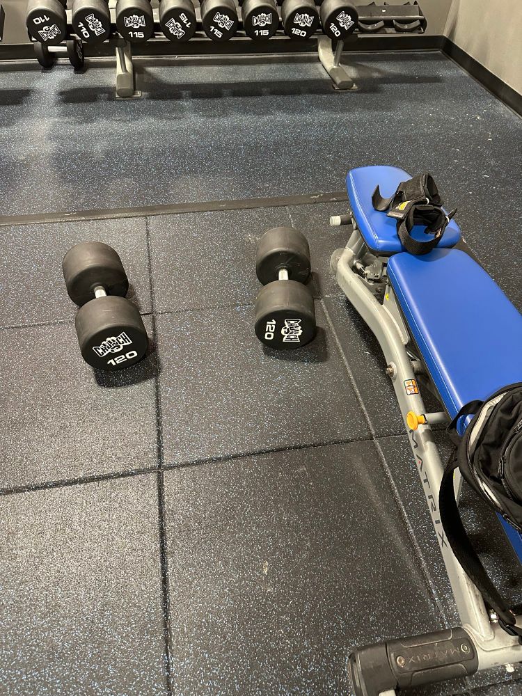 Two 120# dumbbells next to a bench with a backpack and wrist straps next to it 