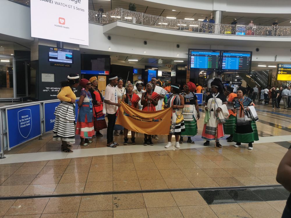 A group of people in a variety of traditional South African clothes, singing and dancing. 