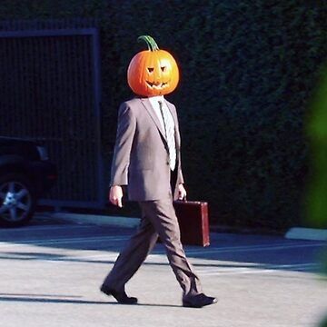 A man wearing a suit and a jack o' lantern on his head walks through a parking lot carrying a briefcase. (Scene from The Office)