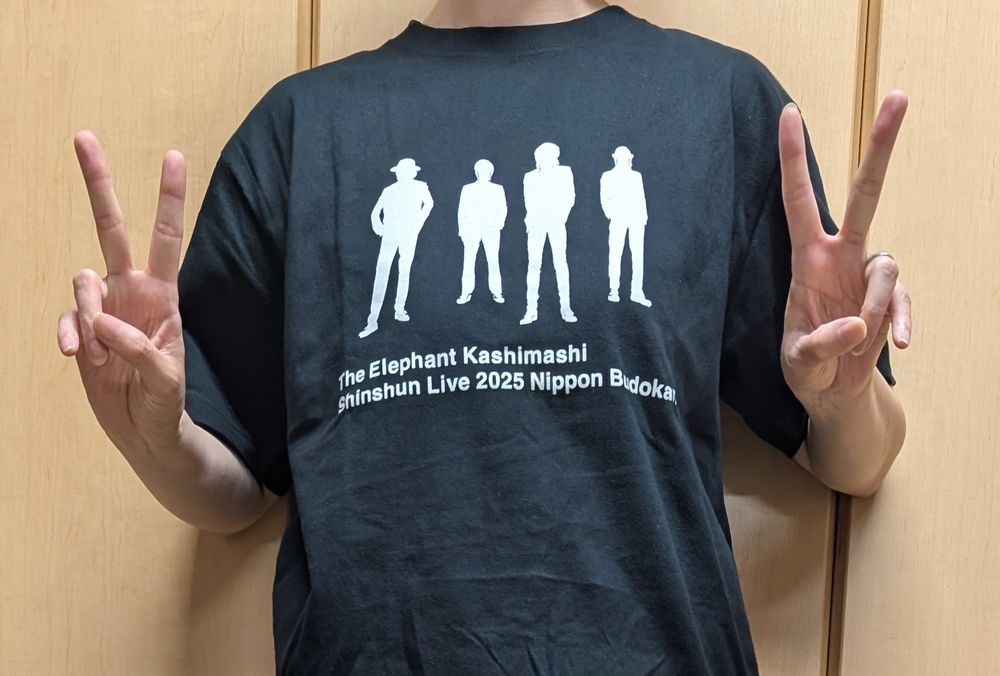 屋内にて、エレファントカシマシのTシャツを着用し、ダブルピースで帰宅報告をする女性の写真です