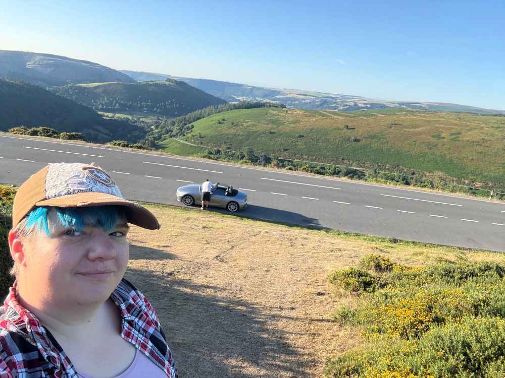 Woman wearing a baseball cap in the foreground looking at the camera. Further down hill behind her a person is facing away from the camera cleaning a convertible's windscreen. The view beyond down the valley is from Horseshoe Pass down the valley to the south.
