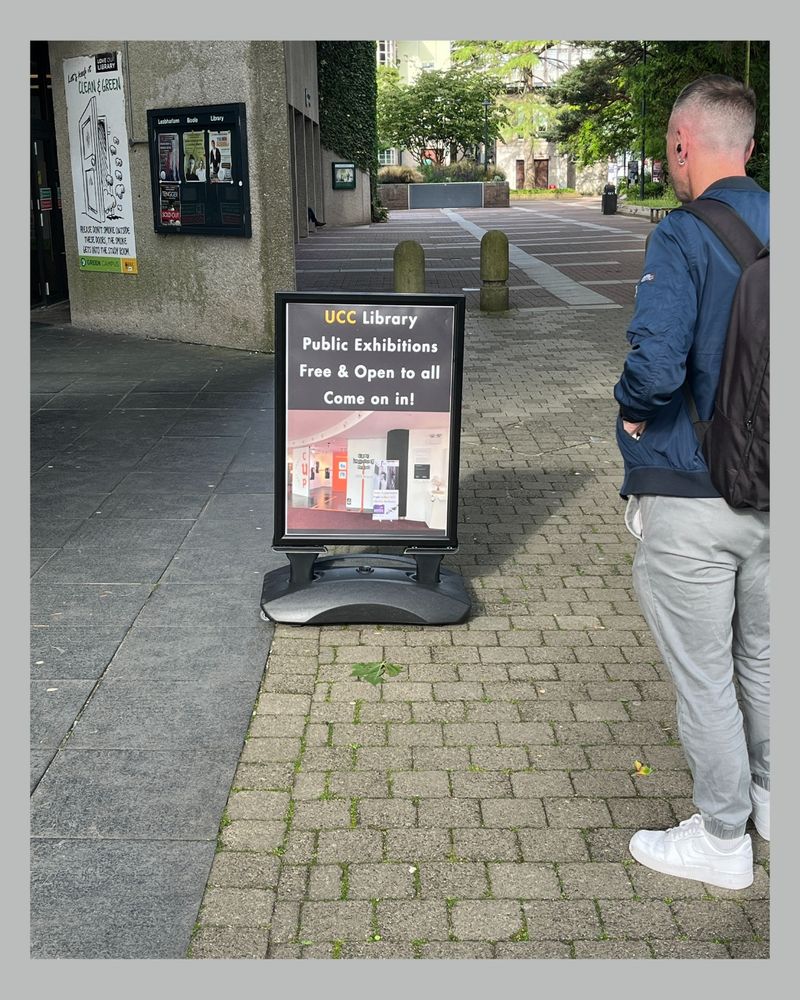 A person with short hair and a backpack stands facing a sign outside UCC Library. The sign reads: "UCC Library Public Exhibitions – Free & Open to all – Come on in!" The library entrance and posters are visible in the background.