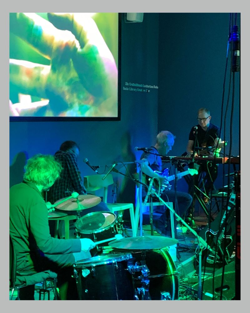 A band of four musicians performs in a dimly lit room with blue and green lighting. The drummer is in the foreground, two guitarists are seated in the middle, and a keyboardist stands on the right. A large screen in the background displays an abstract image of hands.