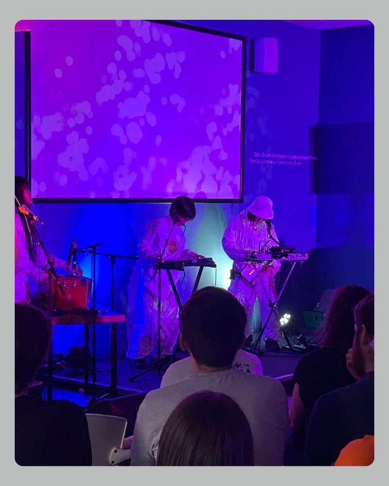 A live performance in a dimly lit room with purple and blue lighting. Three musicians on stage play electronic instruments while wearing shiny, metallic outfits. The audience is seated and watching attentively. A large screen behind the musicians displays abstract patterns