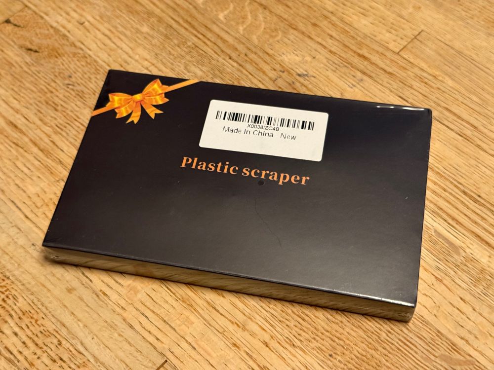 A small black box that says “Plastic Scraper” on it, with a yellow/gold ribbon in the upper left corner like it’s a box of chocolates