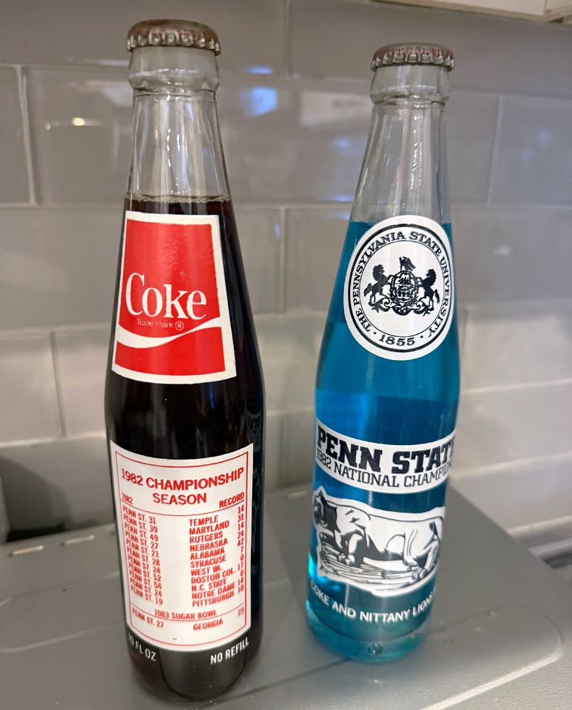 Two 1982 Penn State national championship sodas. Both in glass bottles, sealed, and full. One is the front, it has the school seal, a school logo (a statue of a lion), and PENN STATE 1982 NATIONAL CHAMPIONS on the front. It also says "Coke And Nittany Lions #1!"

The other bottle is full of soda-colored liquid, has the Coke logo, and has a full PSU schedule from 1982 along with NO REFILL and 10 FL OZ.