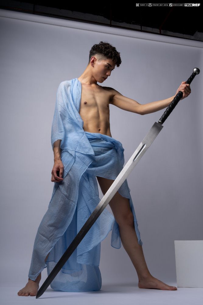 Art pose reference of a young man holding a large sword while draped in light blue fabric. 