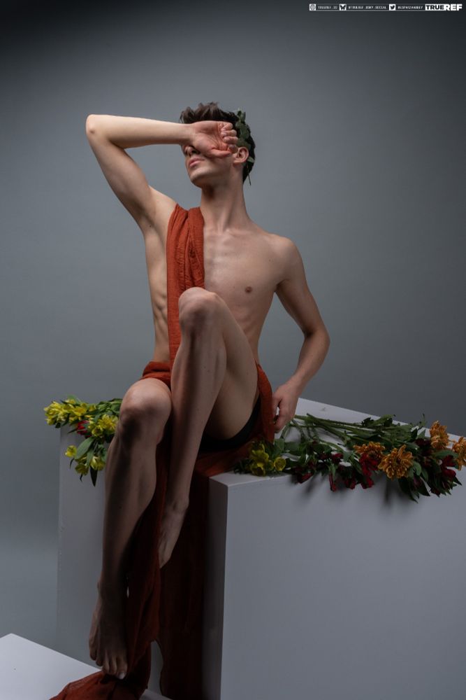 Pose reference of a man draped in red fabric, sitting on a box surrounded by flowers.