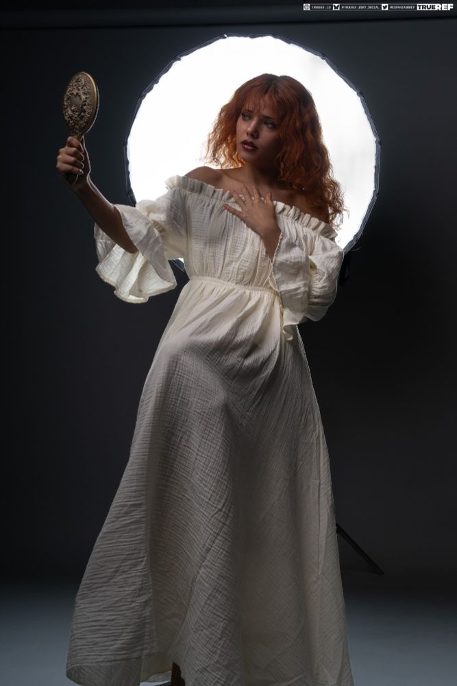 A white woman with red hair and freckles sits in front of a large backlight. She's wearing a white ruffled gown while holding up a vintage hand mirror, admiring herself. 