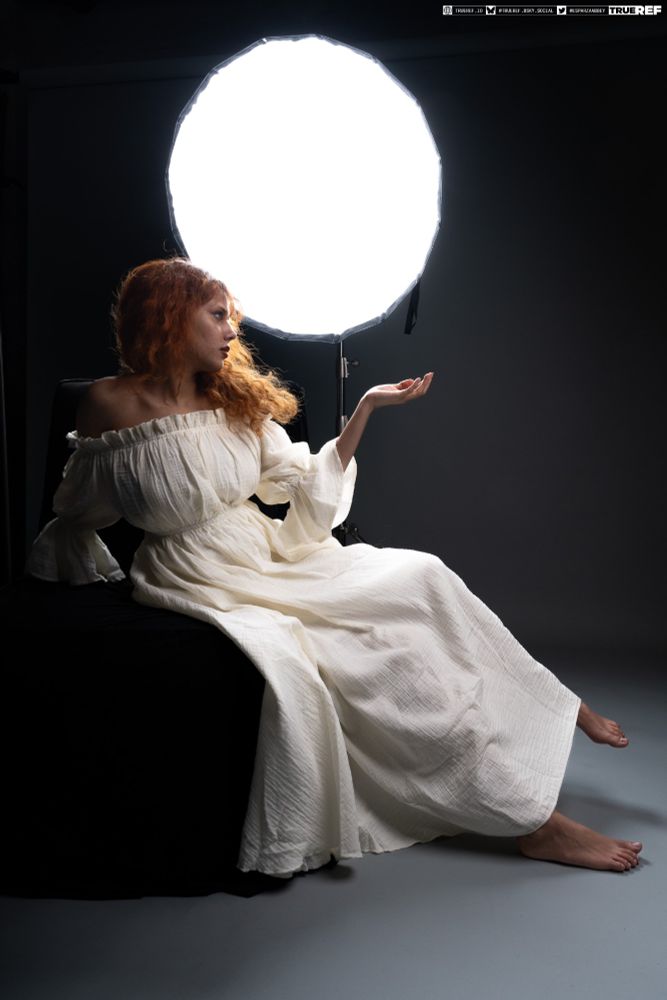 A white woman with red hair and freckles sits on a black chair in front of a large backlight. She's wearing a white ruffled gown while holding her hand out.