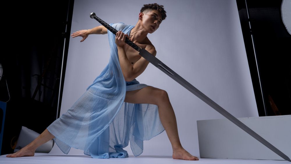 art pose reference of a young man draped in light blue fabric holding a sword. 