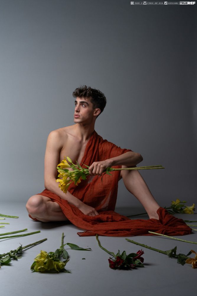 Pose reference of a man draped in red fabric, sitting surrounded by flowers.