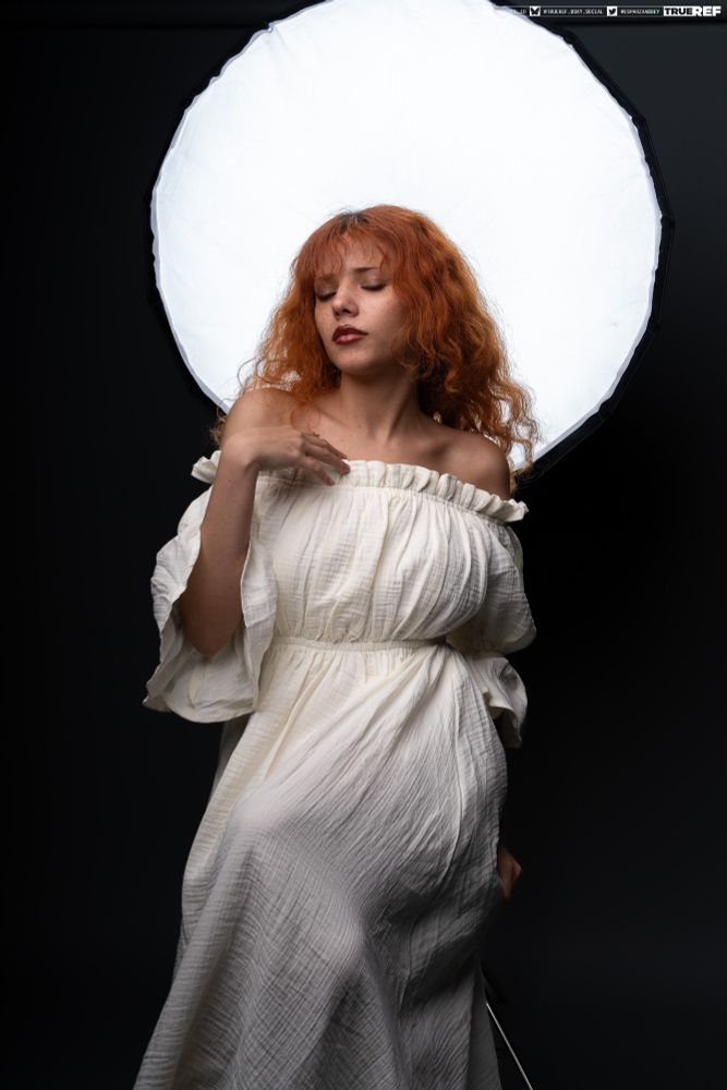 A white woman with red hair and freckles sits in front of a large backlight. She's wearing a white ruffled gown.