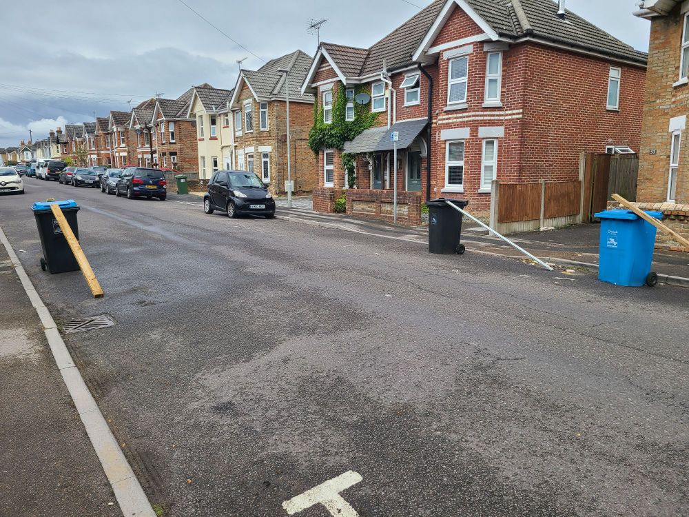 A picture of a street with 3 wheelie bins all with long thin pieces of wood propped up on them like ramps