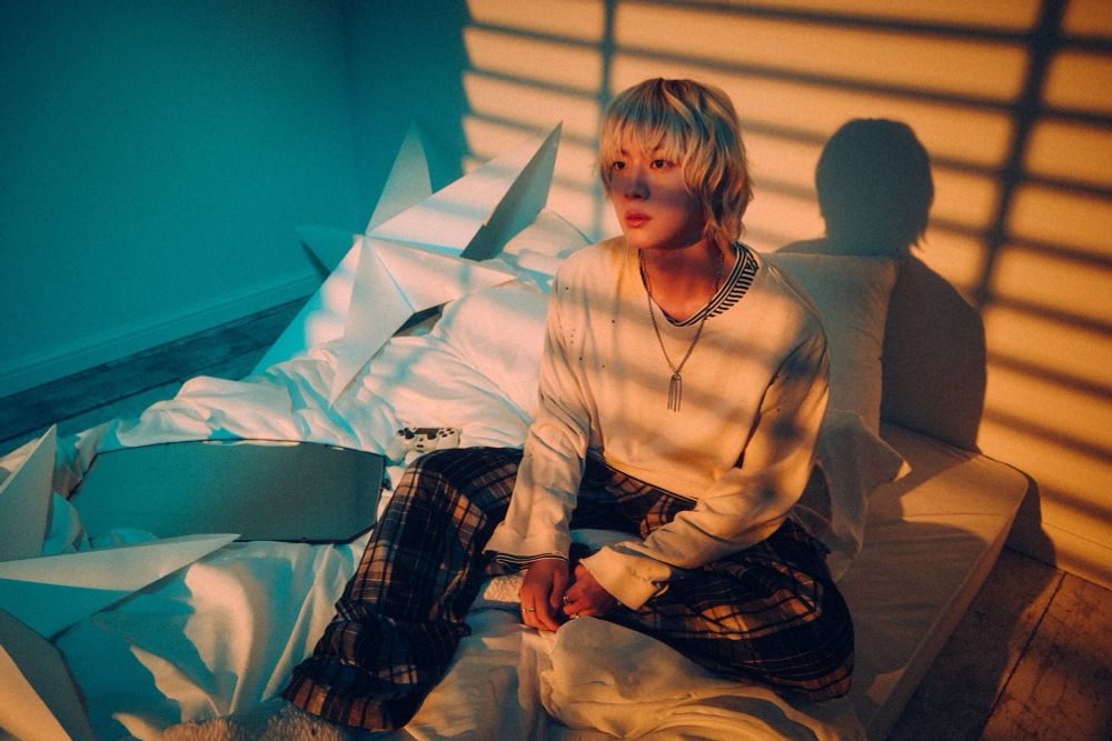 Blonde seokjin sitting on a mattress on the ground surrounded by pillows. A yellow light filtering through from the blinds. 