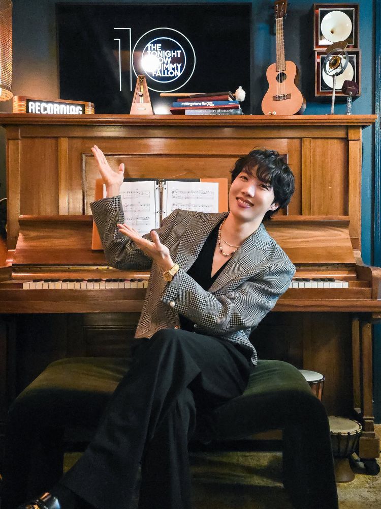 Hobi sitting by the piano crossed-legs and beaming