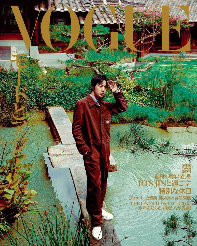 Jin in coordinated maroon / brown-ish outfit standing on a ledge by a body of water